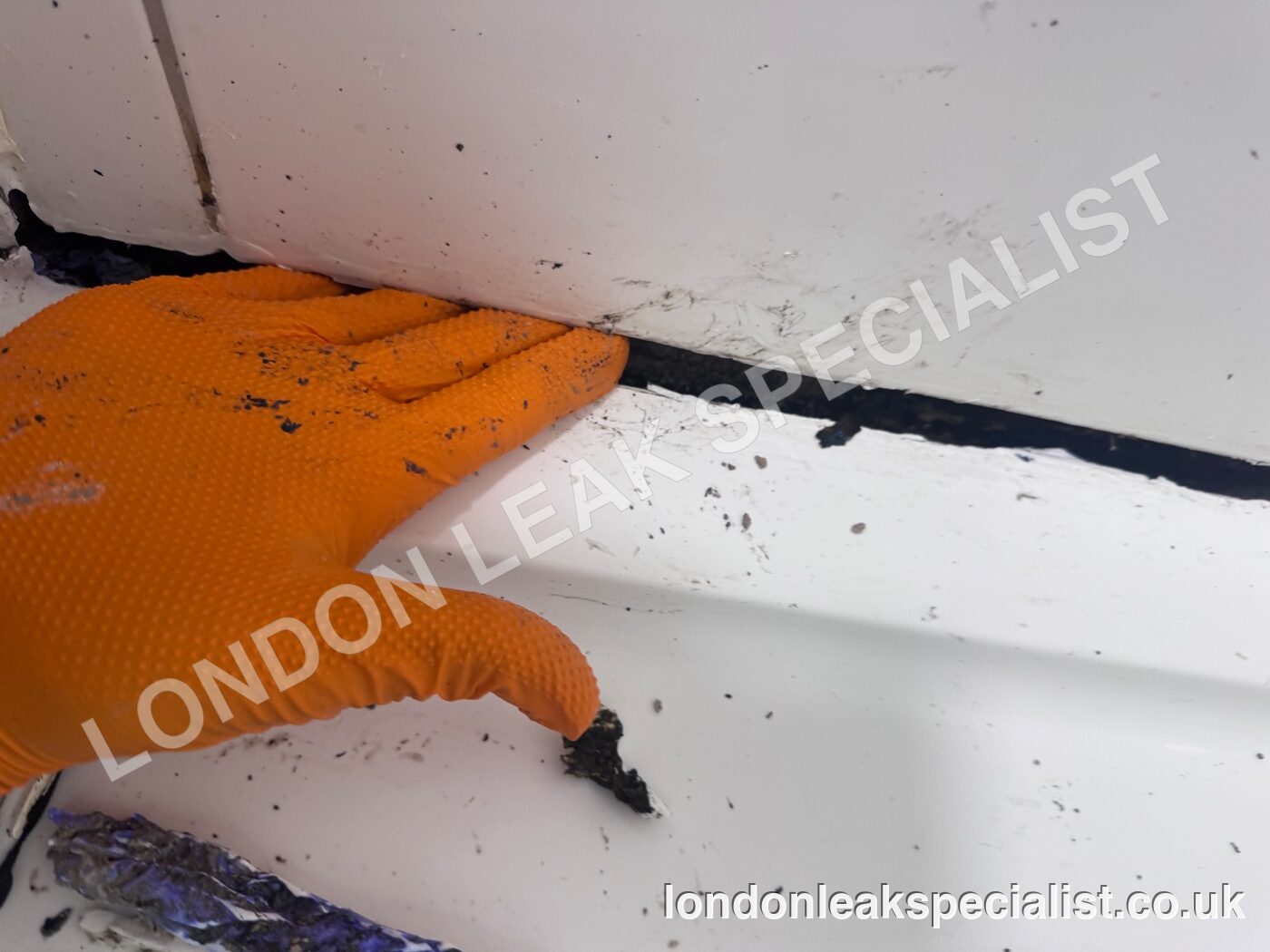A gloved hand pointing to a dark gap where the sealant has failed along the edge of a shower tray