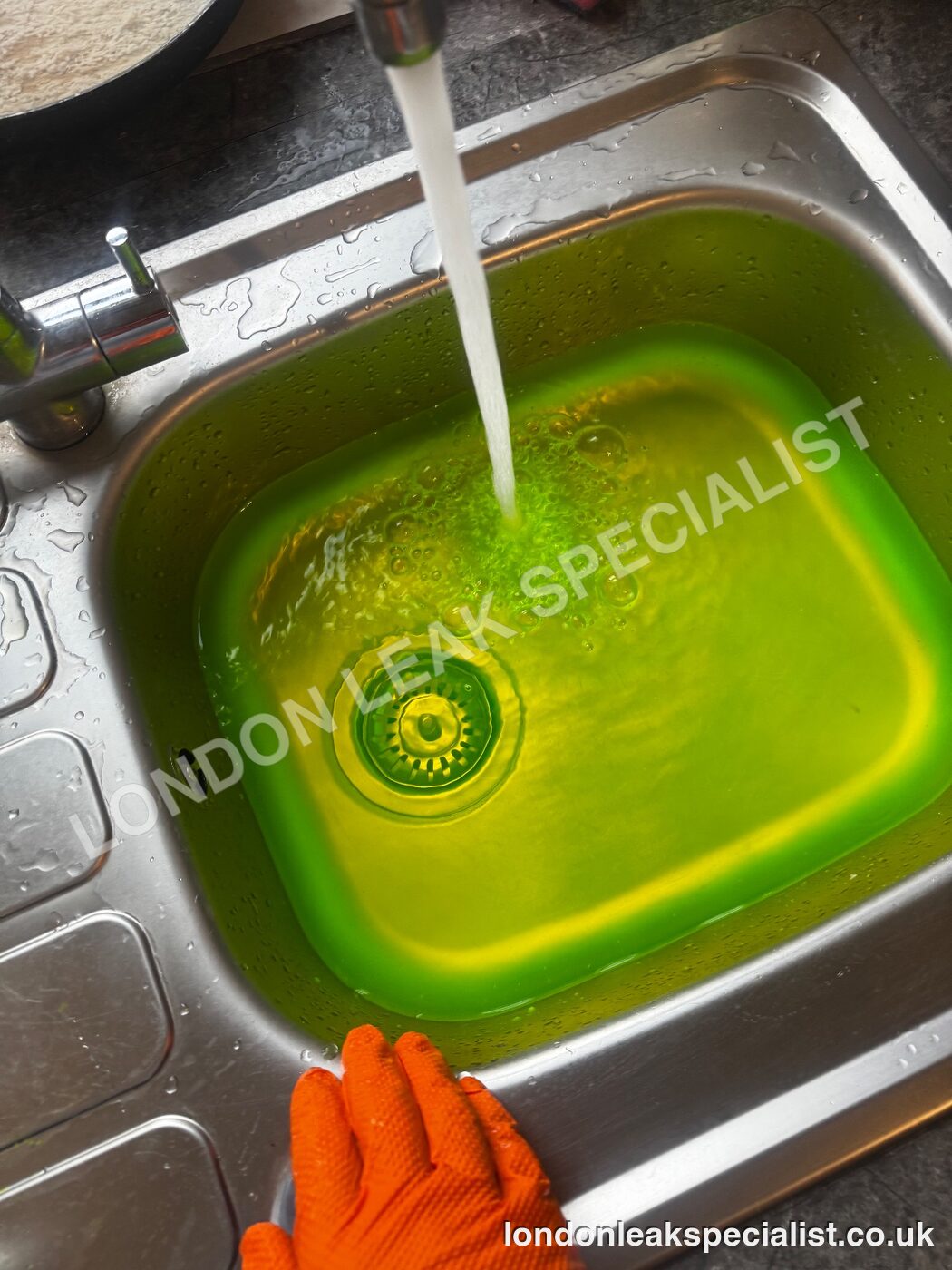 A stainless steel kitchen sink filled with bright green tracer dye water, a gloved hand at the edge
