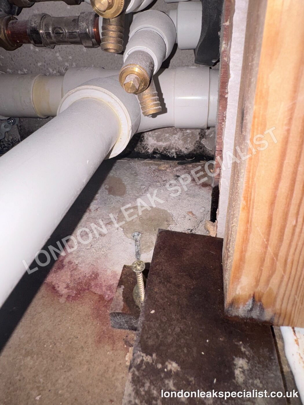 White waste pipework and brass valves in a void beside a bath, with a stained floor surface below