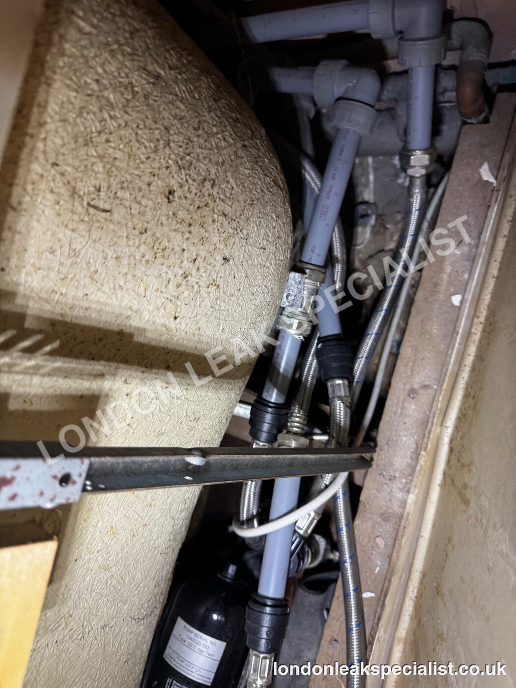 Grey waste pipes and copper supply pipes behind a removed bath panel, with a tool resting across the opening